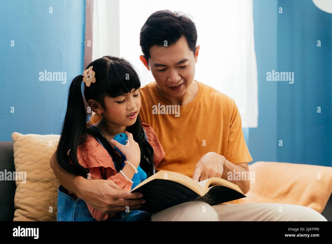 Asiatischer Vater umarmt Tochter und liest Märchenbuch, während er zu Hause auf dem Sofa in der Nähe des Fensters sitzt Stockfoto