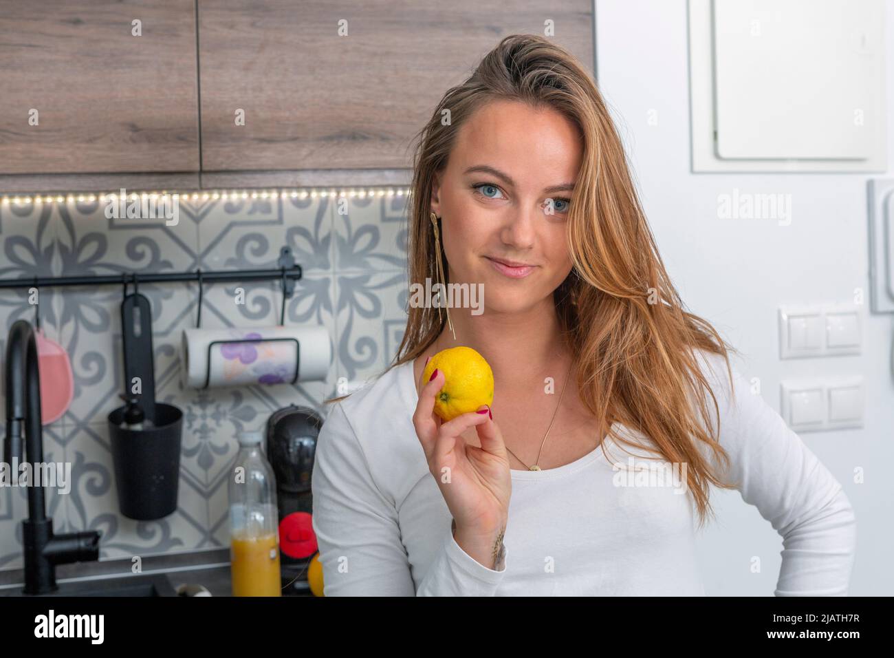Schöne junge Blondine mit blauen Augen in der Küche, die eine Zitrone in der Hand hält und direkt auf die Kamera blickt Stockfoto