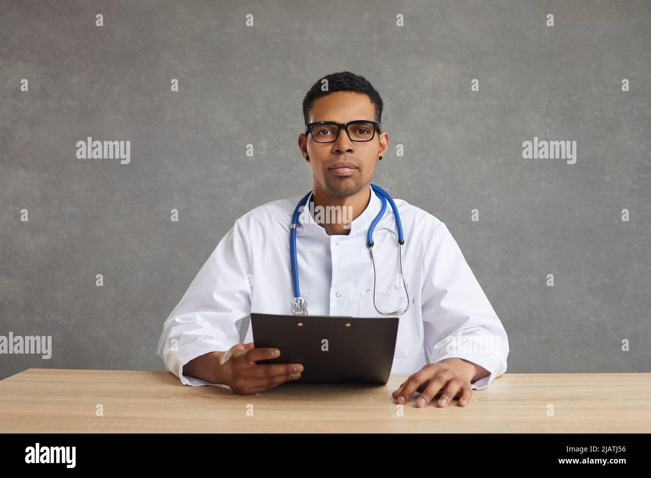 Seriöse afroamerikanische Ärztin, die auf die Kamera schaut, die auf dem Schreibtisch im Studio sitzt Stockfoto
