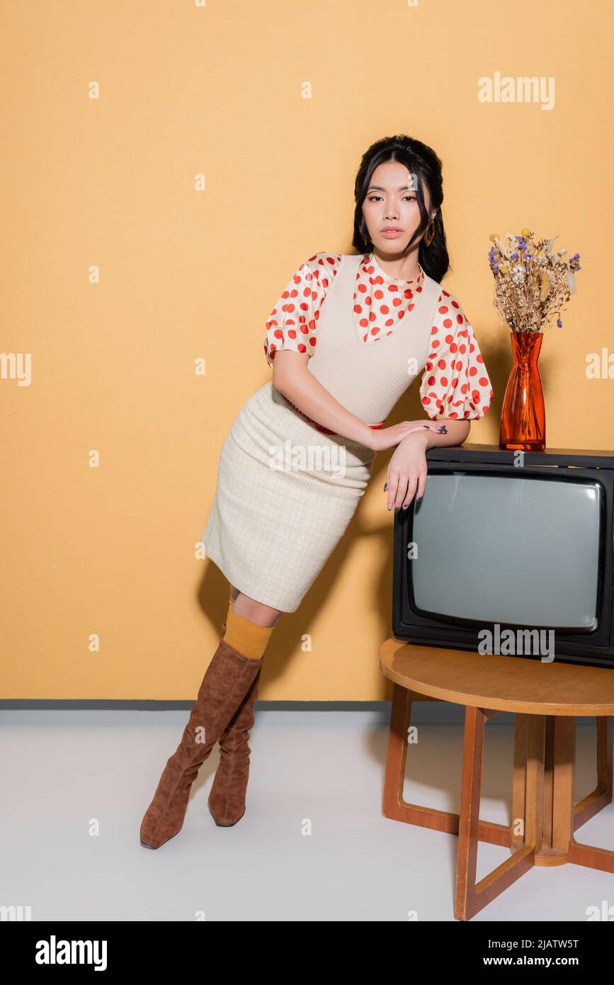 Stilvolle asiatische Frau Blick auf Kamera in der Nähe von Retro-tv und Blumen auf orangefarbenem Hintergrund Stockfoto