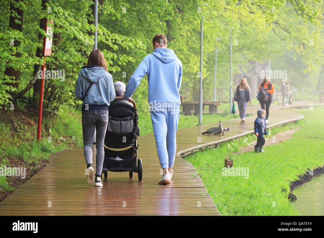 Junges Paar mit Kinderwagen, das nach dem Regen in einem Park spazierengeht. Familienurlaub an der Seeufer im Sommer Stockfoto