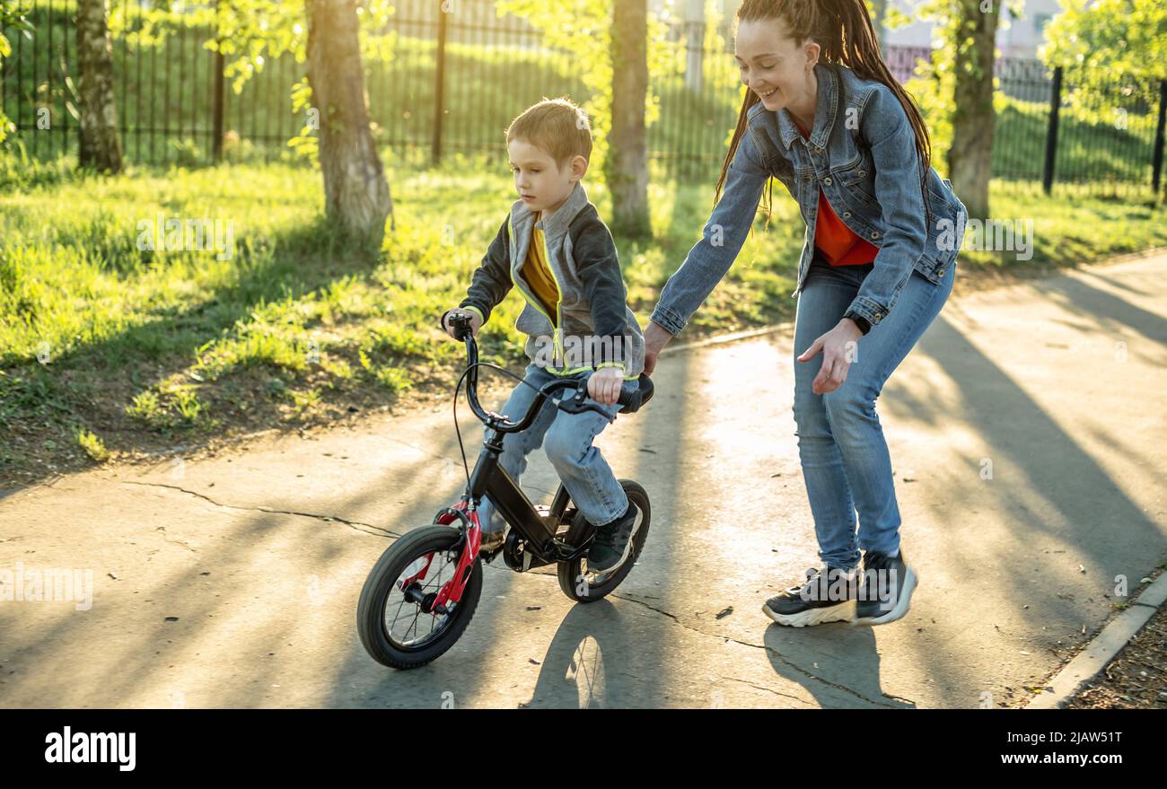 Mama hilft einem kleinen Jungen, ein Fahrrad mit zwei Rädern im Park zu fahren. Ein angenehmer Sommersporturlaub für Kinder. Stockfoto