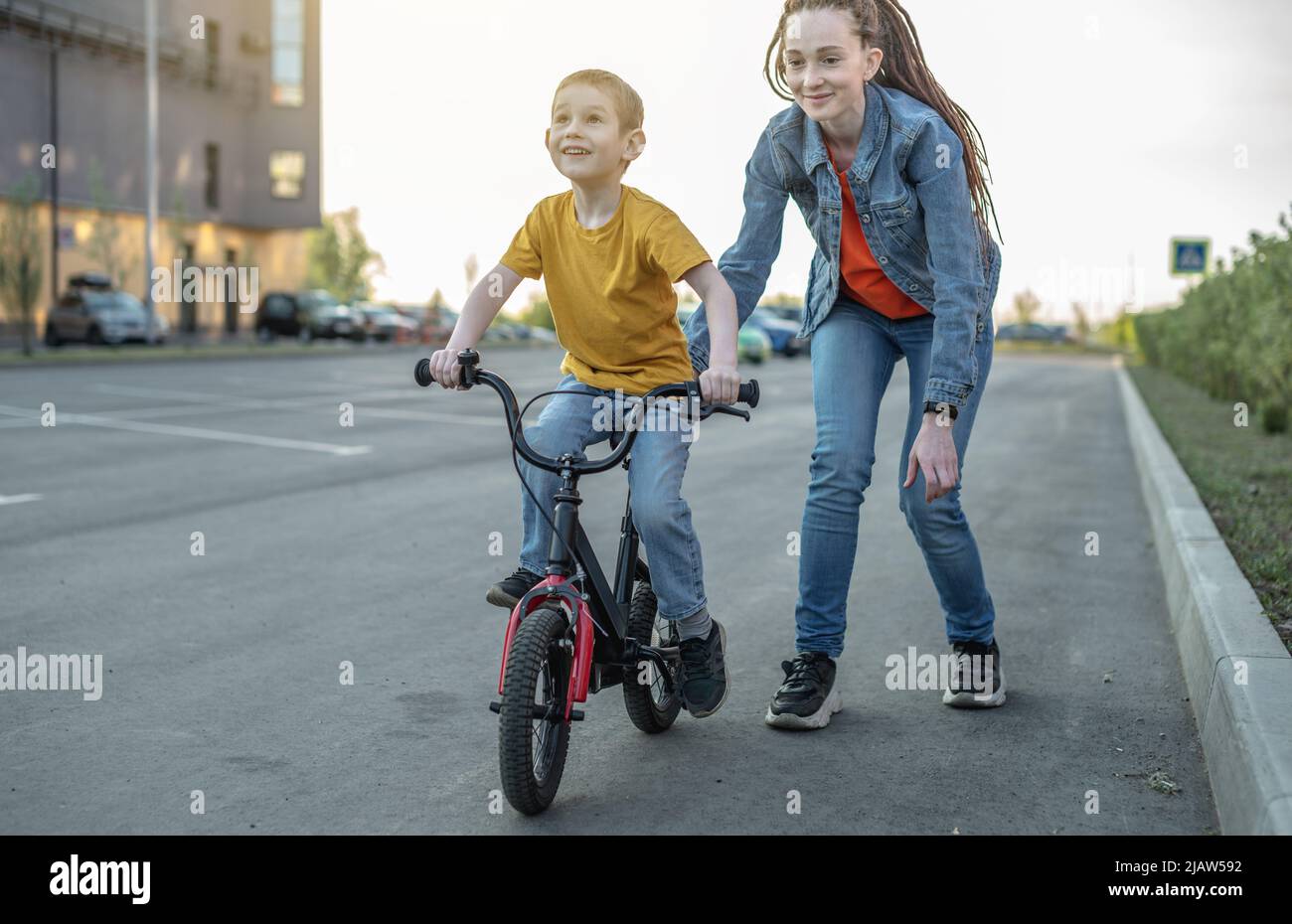 Mama hilft einem kleinen Jungen, ein Fahrrad mit zwei Rädern im Park zu fahren. Ein angenehmer Sommersporturlaub für Kinder. Stockfoto