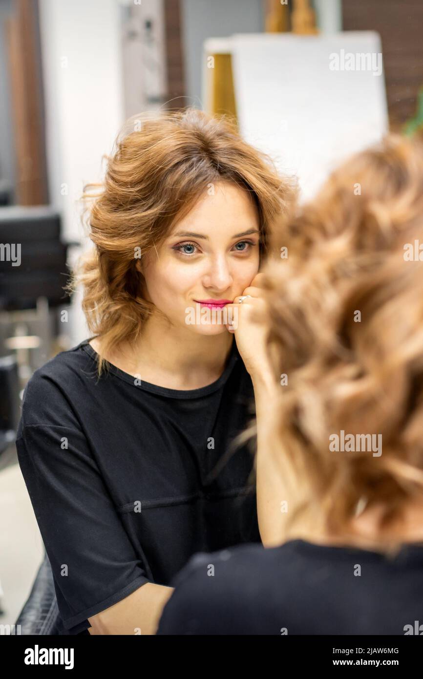 Die schöne junge kaukasische rothaarige Frau mit einer neuen kurzen welligen Frisur, die im Spiegel aussieht und Frisur und Make-up in einem Friseursalon überprüft Stockfoto