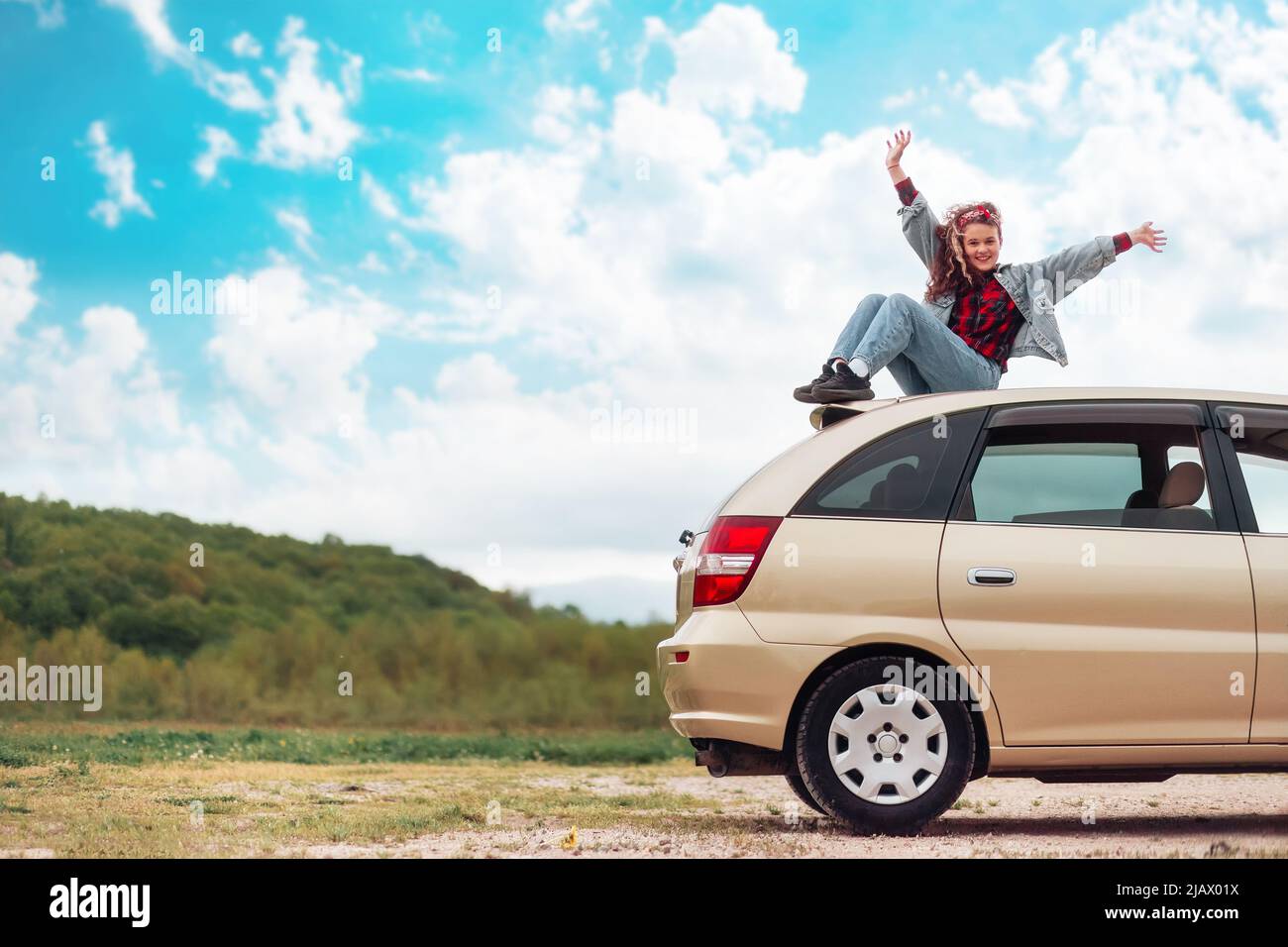 Autofahrt. Glückliches Teenager-Mädchen sitzt auf dem Dach eines Autos und hebt freudig ihre Hände hoch. Berge und Himmel sind im Hintergrund. Speicherplatz kopieren. Der Nachteil Stockfoto