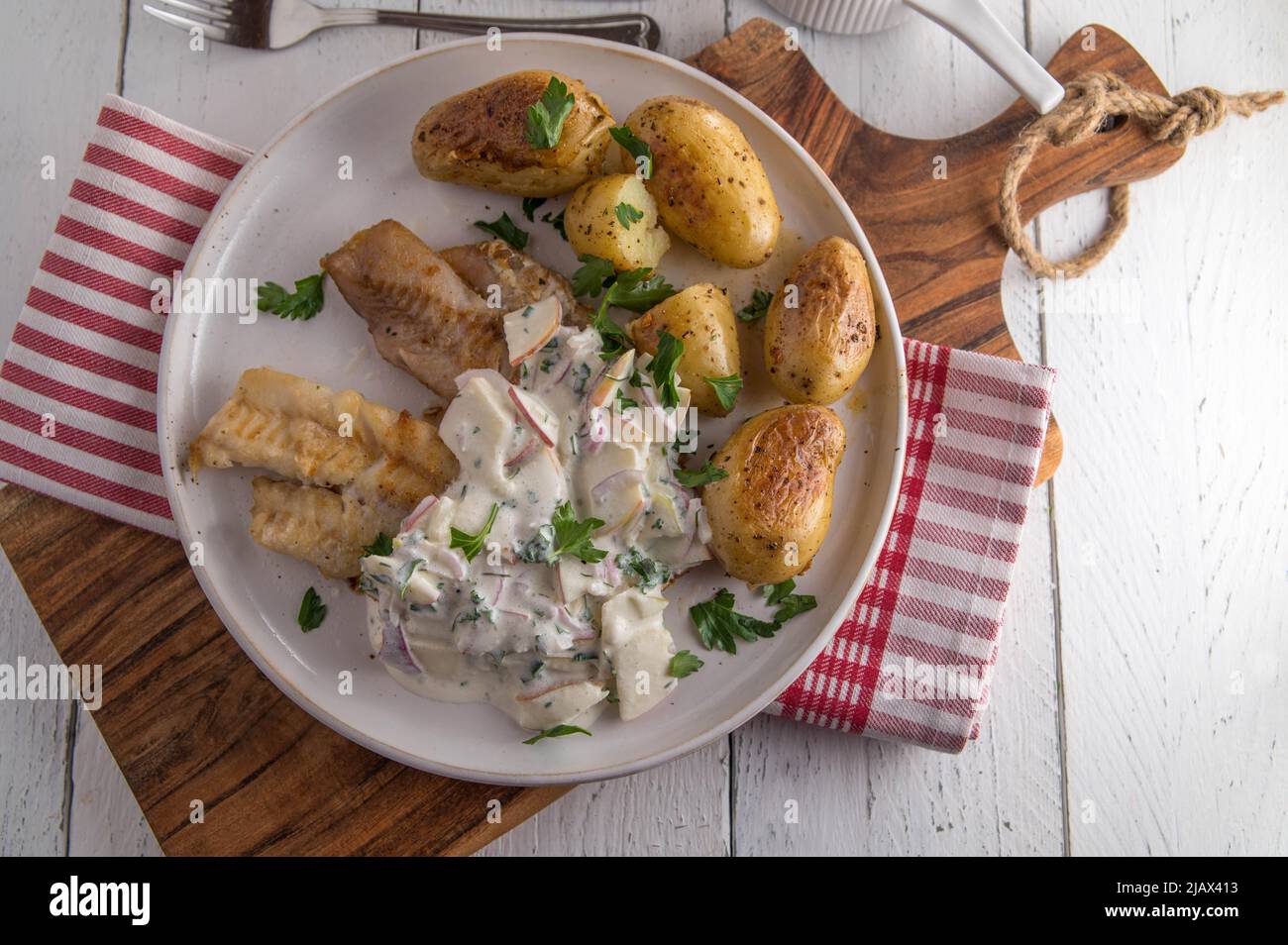 Gebratener Koalfisch mit Sauerrahm, Apfel, Zwiebel, Kräutersauce. Serviert mit gebratenen Kartoffeln auf einem Teller isoliert auf weißem Holztisch Hintergrund Stockfoto
