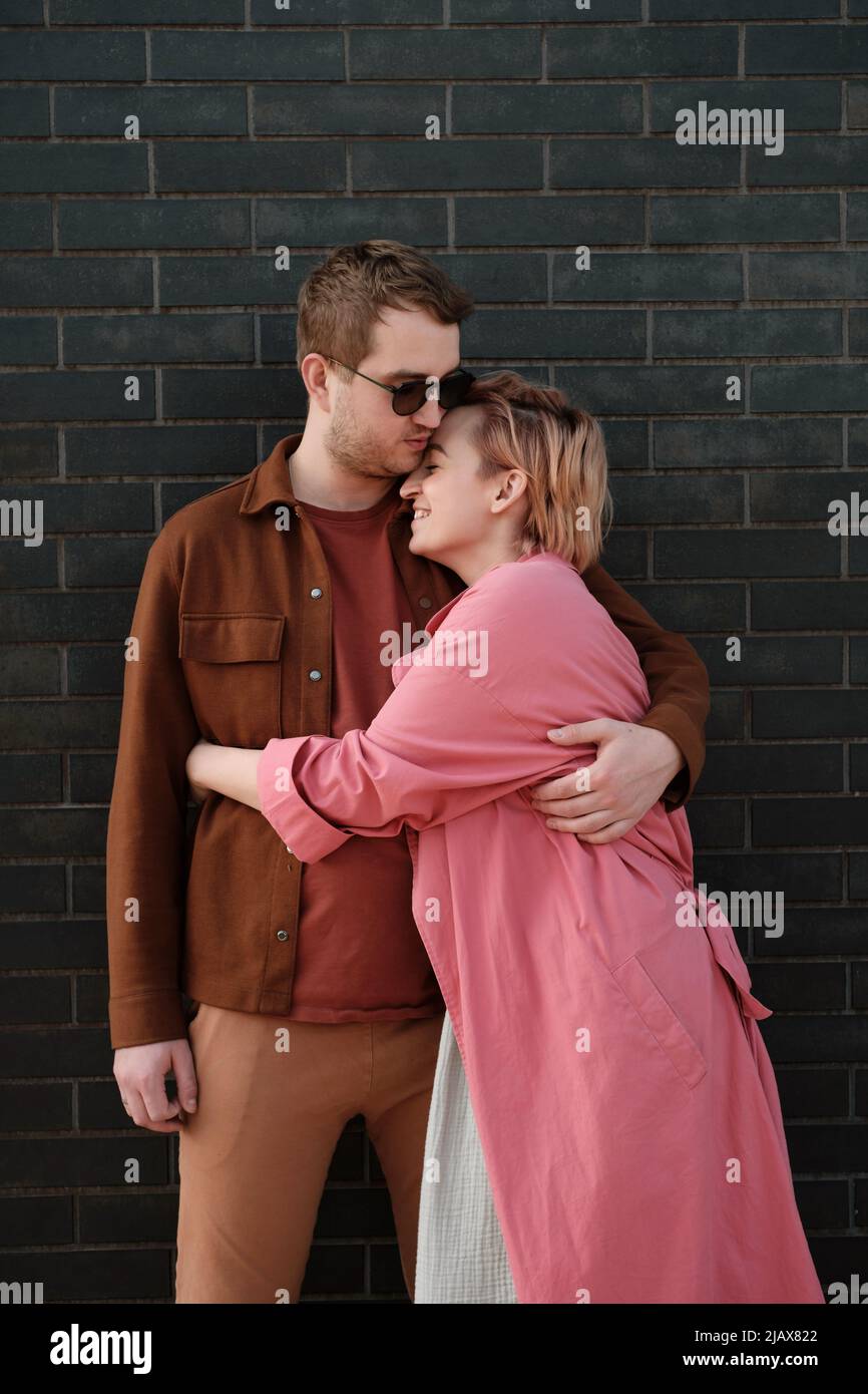 Pärchen umarmt sich über die dunkle Wand. Glücklicher Mann und Frau berühren sich zärtlich. Liebe und Romantik. Stylische Menschen in Pink und Braun kuscheln sich Stockfoto