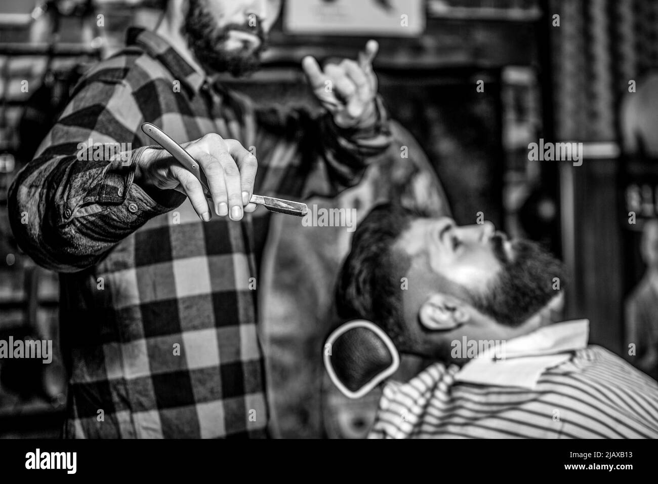 Männer Barbier rasiert seinen Bart. Bärtiger Mann sitzt in einem Sessel in einem Friseurladen, während Friseur seinen Bart mit einem gefährlichen Rasiermesser rasiert. Friseur Stockfoto
