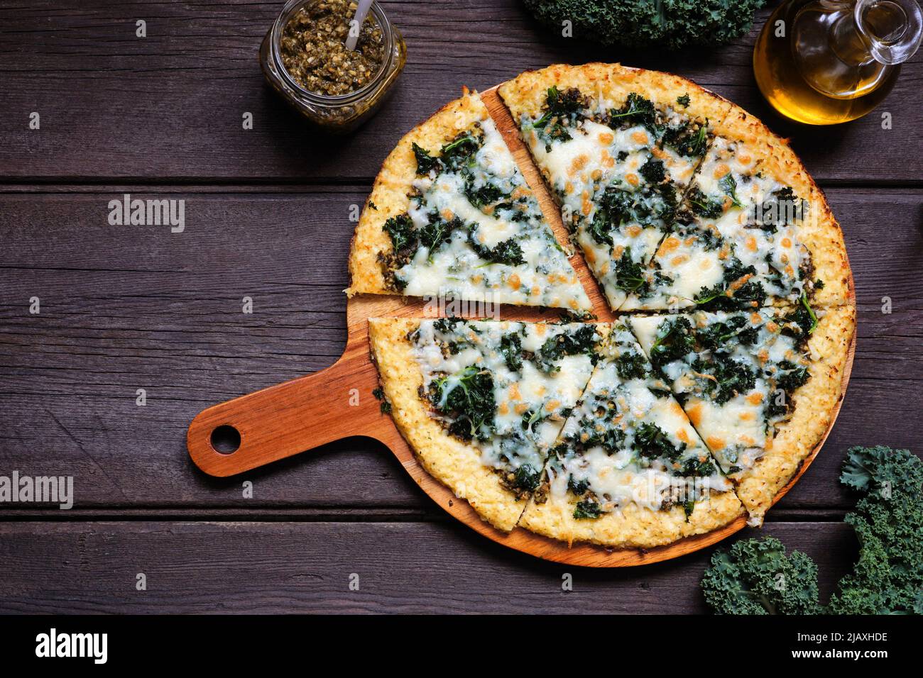 Gesunde, kohlenhydratarme Blumenkohlkruste Pizza mit Grünkohl und Pesto-Sauce. Obere Ansicht mit geschnittenen Schichten. Tischszene auf dunklem Holzhintergrund. Stockfoto