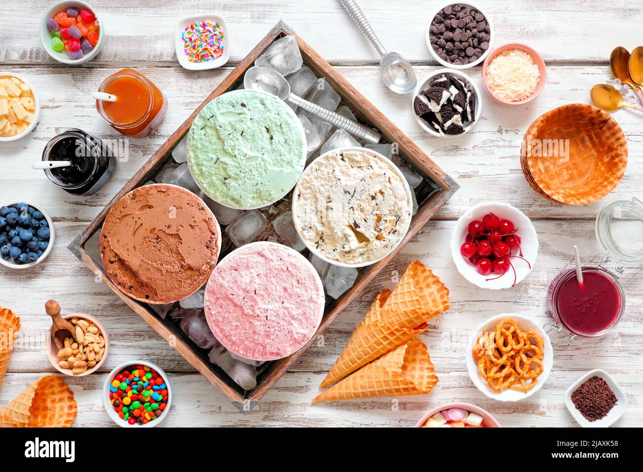 Sommerliches Eisbuffet mit verschiedenen Eisaromen und süßen Belägen. Deckenansicht Tischszene auf einem rustikalen weißen Holzhintergrund. Stockfoto