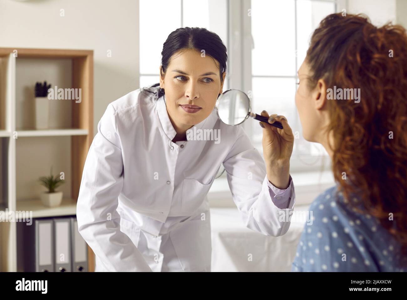 Professioneller Dermatologe mit Lupe, um die Haut auf dem Gesicht der Frau zu untersuchen Stockfoto