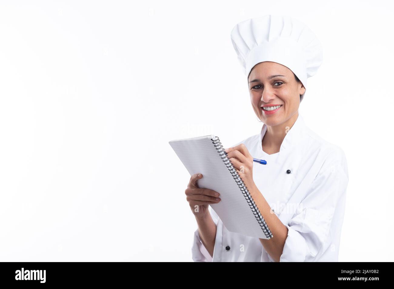 Glückliche Latina-Köchin schreibt Rezepte in ihr Notizbuch und schaut auf die Kamera isoliert auf weißem Hintergrund mit Kopierplatz. Stockfoto