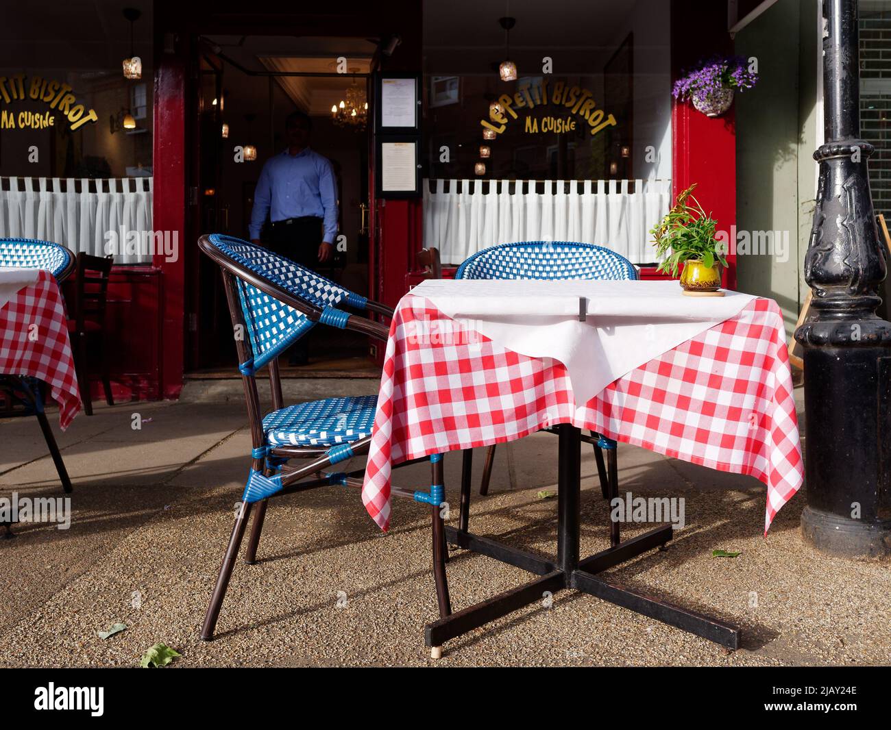 MA Cuisine La Petit Bistro im Dorf Kew. Kellner in einem blauen Hemd mit Schatten verdecktem Kopf nähert sich den Tischen draußen. London. Stockfoto