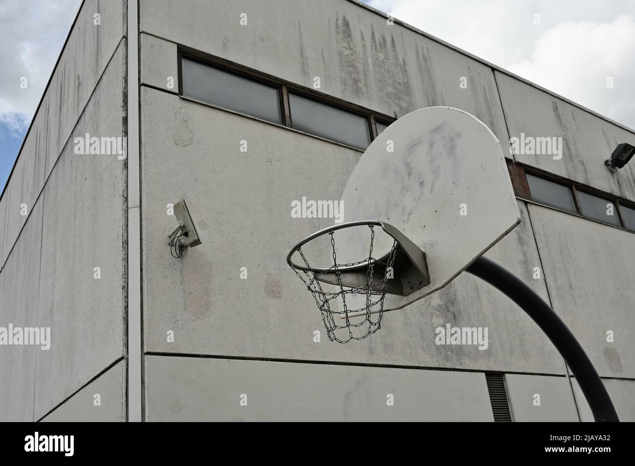 Ein vandalismussicherer Basketballkorb mit eisernen Ketten vor einem düsteren bewölkten Himmel, neben einem Betongebäude mit einer Überwachungskamera an der Wand Stockfoto