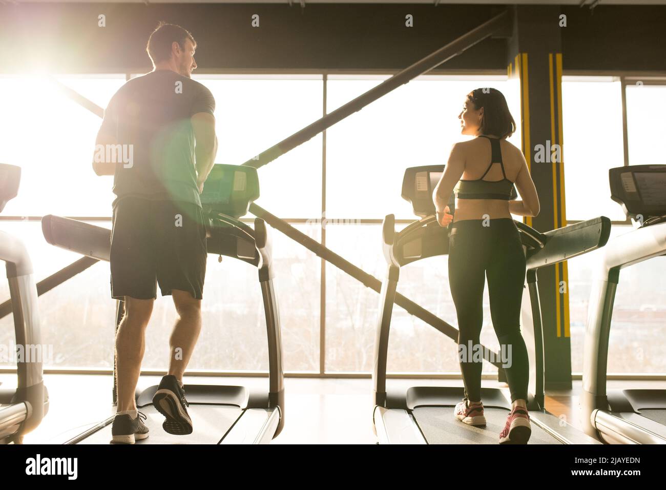 Rückansicht eines jungen Paares, das sich morgens auf Laufbändern vor einem großen Fenster unterhielt Stockfoto