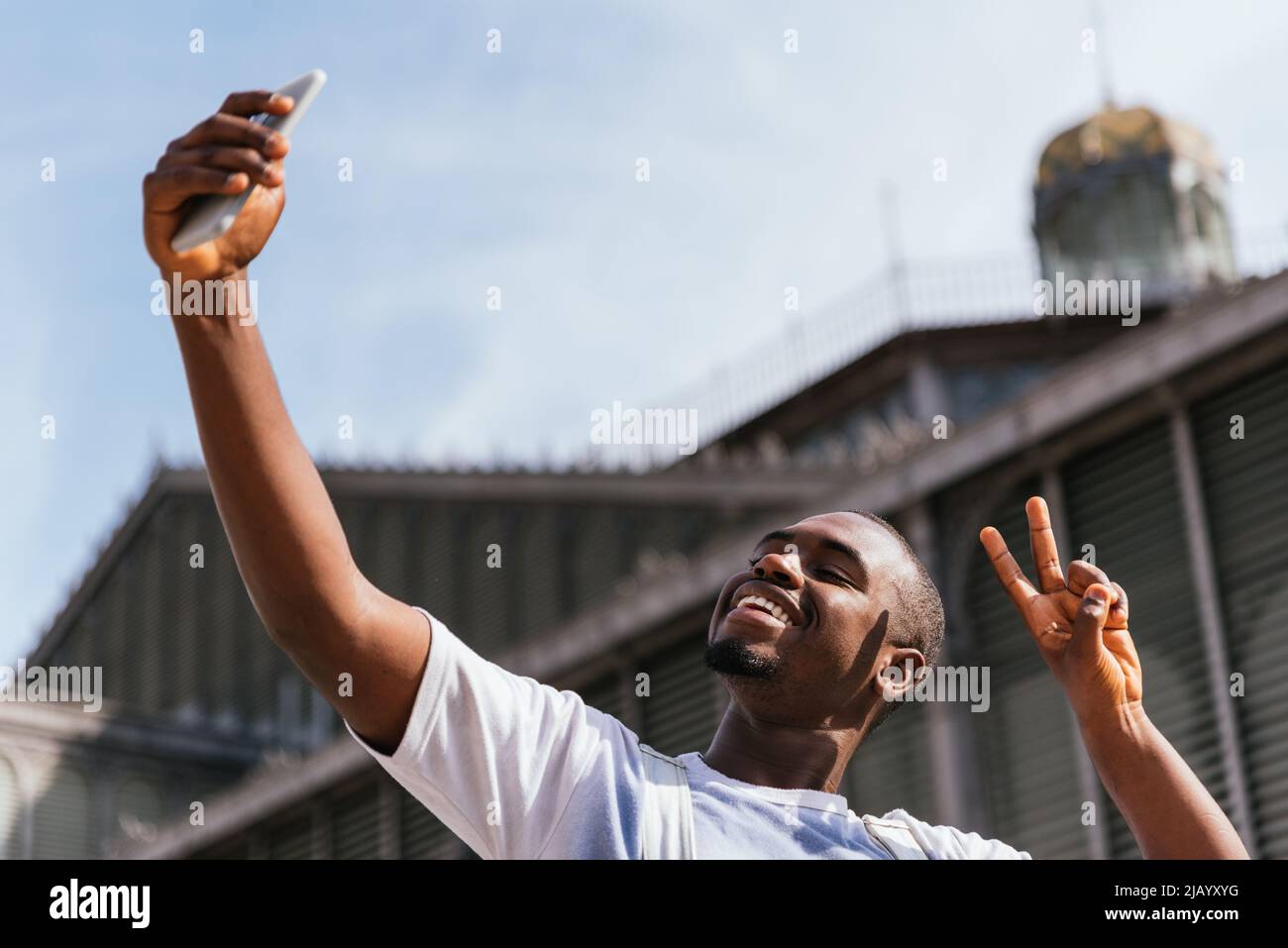 Von unten glücklicher junger afroamerikanischer männlicher Tourist. Er zeigt zwei Finger und lächelt hell, während er am sonnigen Tag Selfie auf dem Smartphone gegen die Kirche macht Stockfoto