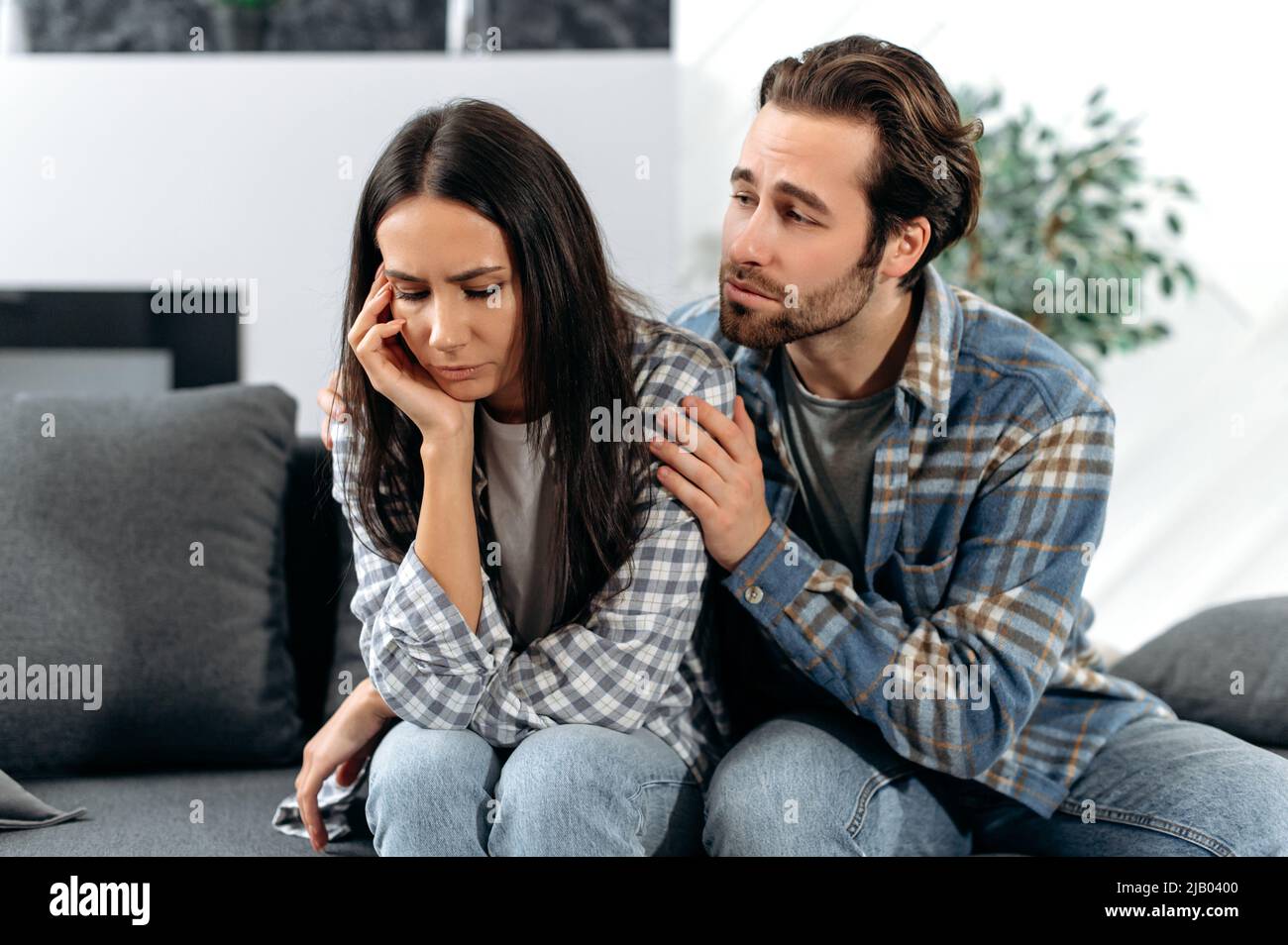 Ein Ehepaar versöhnt sich nach einem Streit. Ein Mann und eine Frau sitzen auf einem Sofa, ein Mann bittet um Vergebung von einer Frau oder tröstet sie, das brünette Mädchen wird von ihrem Mann verärgert oder beleidigt Stockfoto