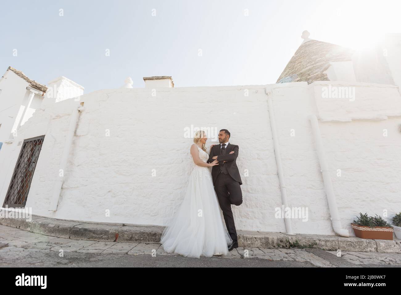 Ein romantisches Ehepaar, eine Frau im weißen Hochzeitskleid und ein Mann im schwarzen Anzug stehen zusammen, schauen sich an der Wand an Stockfoto
