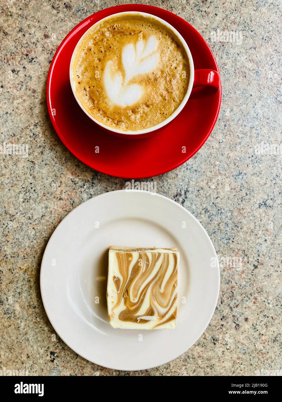 Kaffee und Kuchen. Eine Aufnahme von Latte Art, die in einer roten Kaffeetasse mit einer Kuchenseite präsentiert wird. Stockfoto