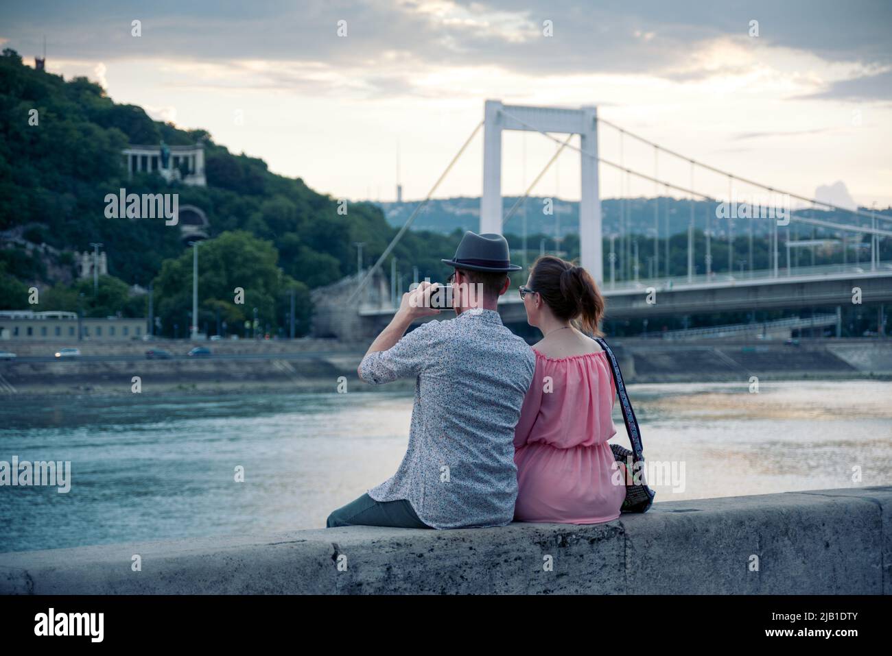 Junges erwachsenes Paar, das am Fluss sitzt und eine Aussicht fotografiert Stockfoto