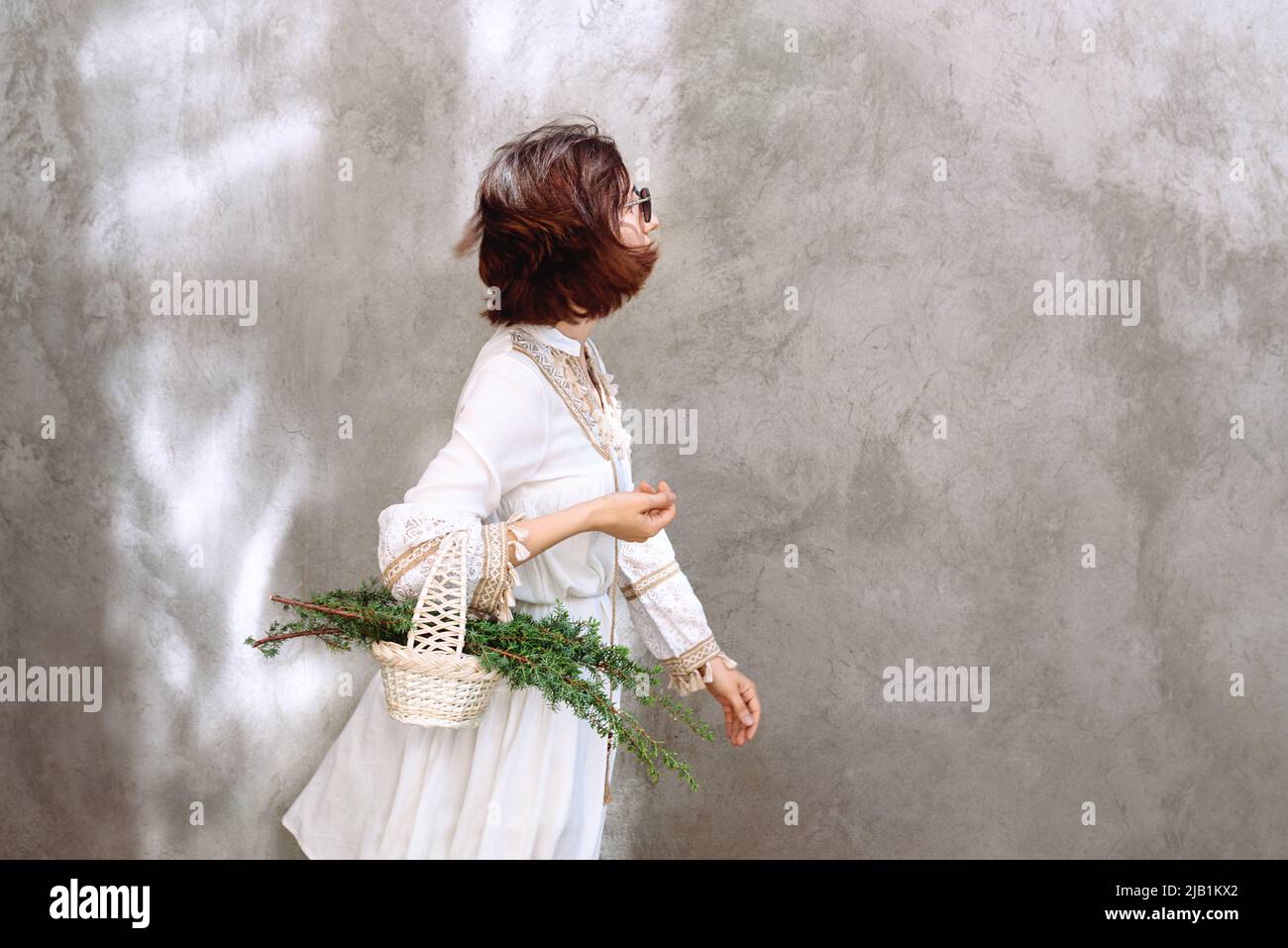 Mädchen ist in Eile und trägt in Korb von Weihnachtsbaum Zweige auf dem Hintergrund der Betonwand mit Sonnenlicht. Frau in weißem Kleid im Boho-Stil. Das c Stockfoto