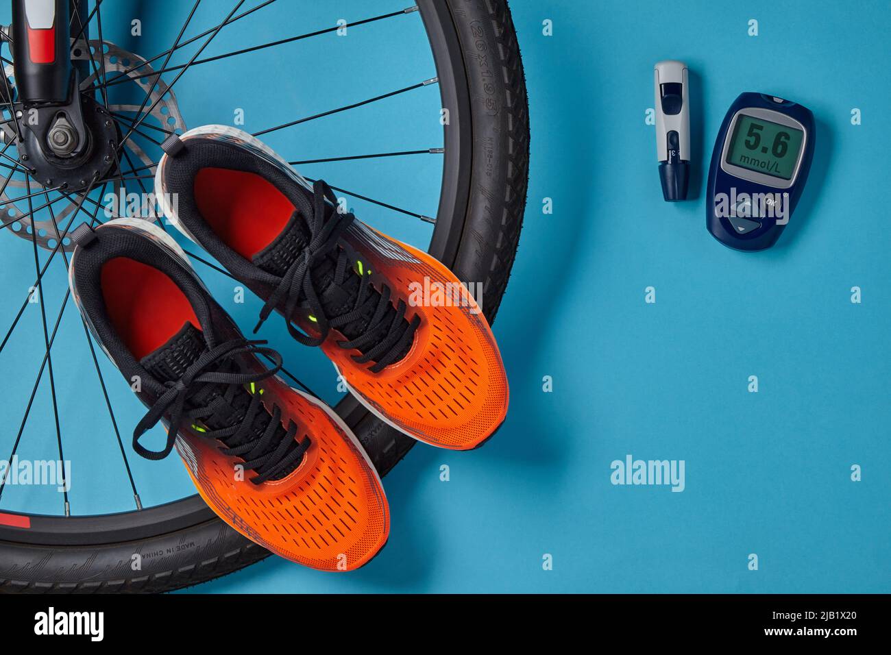 Fahrradrad, orangefarbene Turnschuhe und Blutzuckermessgerät auf blauem Hintergrund. Übung zur Normalisierung des Blutzuckers Stockfoto