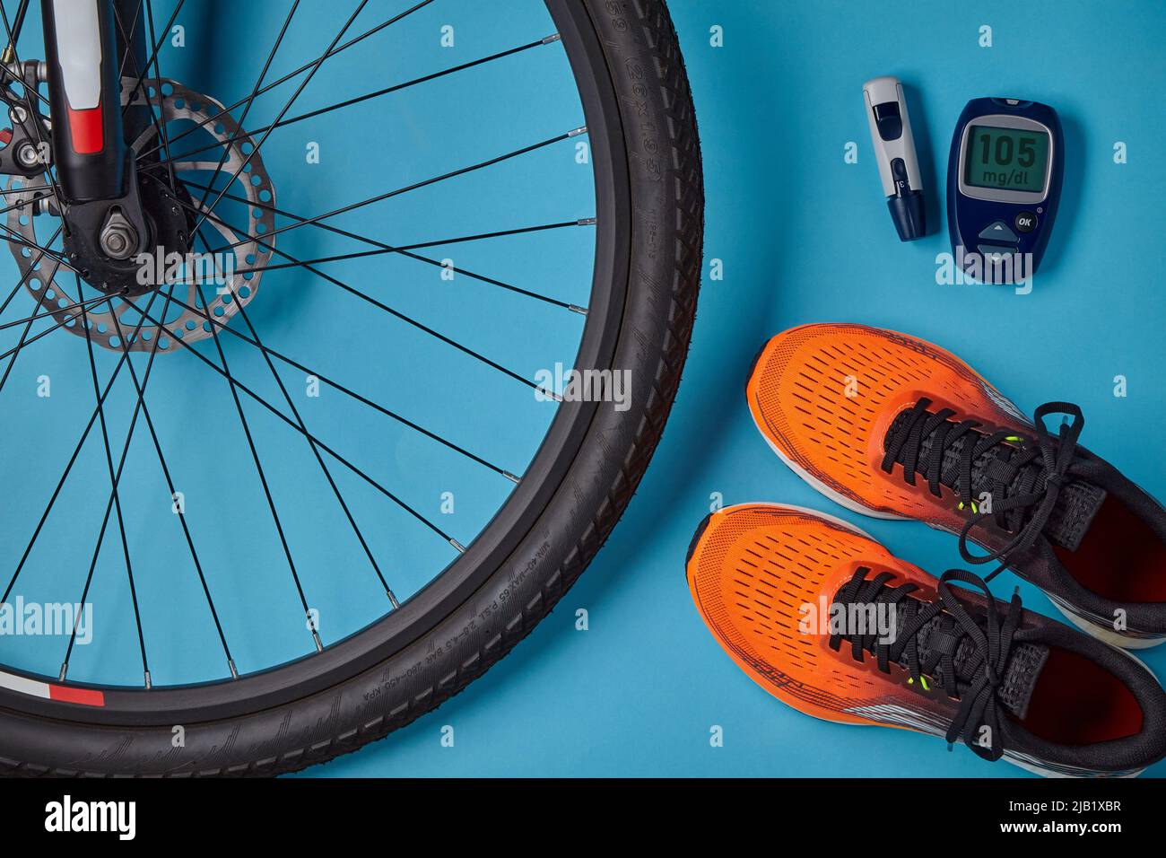 Fahrradrad, orangefarbene Turnschuhe und Blutzuckermessgerät auf blauem Hintergrund. Körperliche Aktivität, um den Blutzucker einer diabetischen Person zu normalisieren Stockfoto