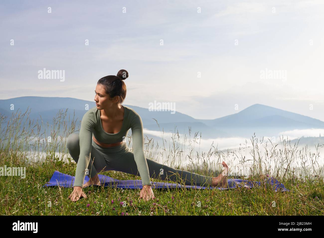 Attraktive Frau mit dunklem Haar, die die Beine auf einer Yogamatte im Freien dehnt. Aktive Dame in Sportkleidung genießt das morgendliche Workout mit Blick auf die Berge. Stockfoto