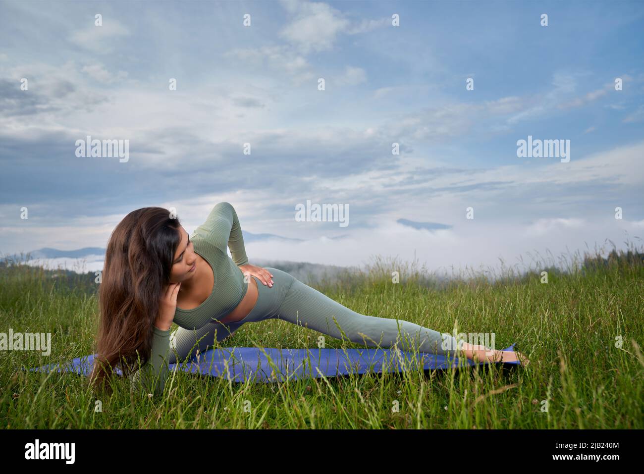Junge Frau mit perfekter Figur genießt Morgenübungen mit Bergen im Hintergrund. Sportliche Brünette trainiert regelmäßig an frischer Luft. Stockfoto