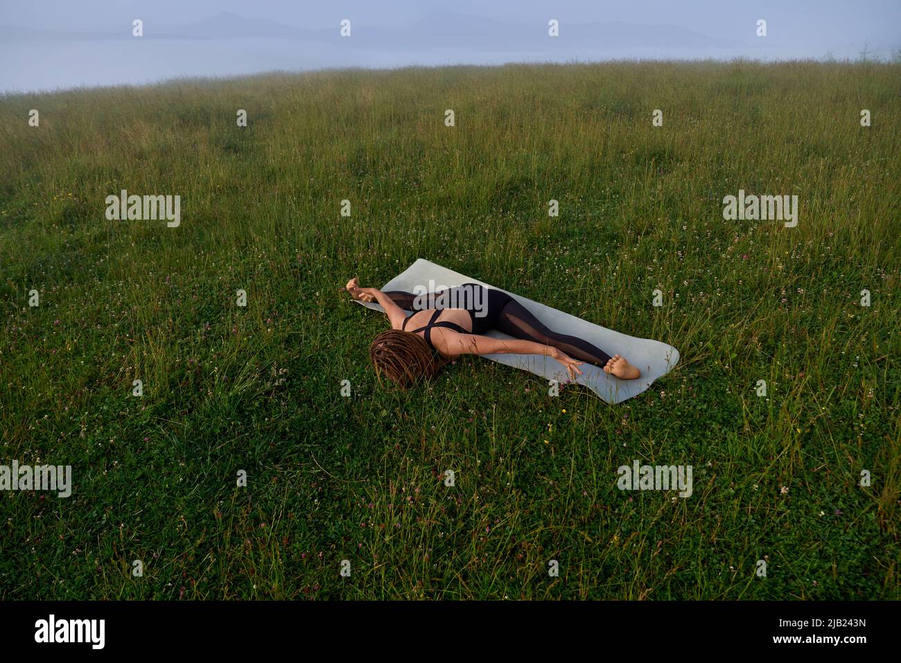 Flexible junge Frau in Sportkleidung Splits auf Yoga-Matte inmitten der grünen Natur. Weibliche Person mit Dreadlocks, die allein an frischer Luft trainiert. Stockfoto