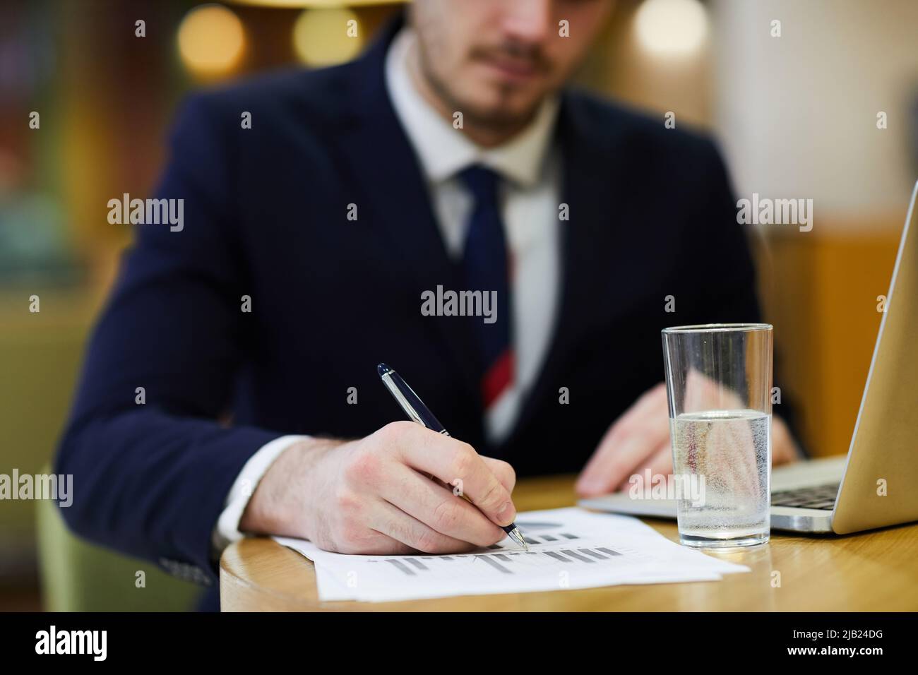 Nahaufnahme eines vielbeschäftigten Analysten, der mit einem Glas Wasser und einem Laptop am Tisch sitzt und mit statistischen Grafiken arbeitet Stockfoto