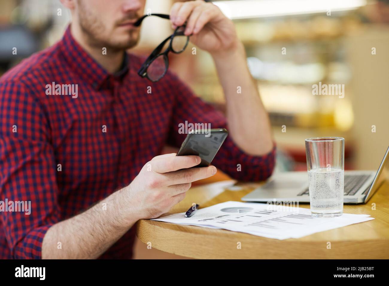 Nahaufnahme eines nachdenklichen, beschäftigten Analysten in legerem Hemd, der mit Finanzbericht, Laptop und Glas Wasser am Tisch sitzt und mit dem Smartphone beißt Stockfoto