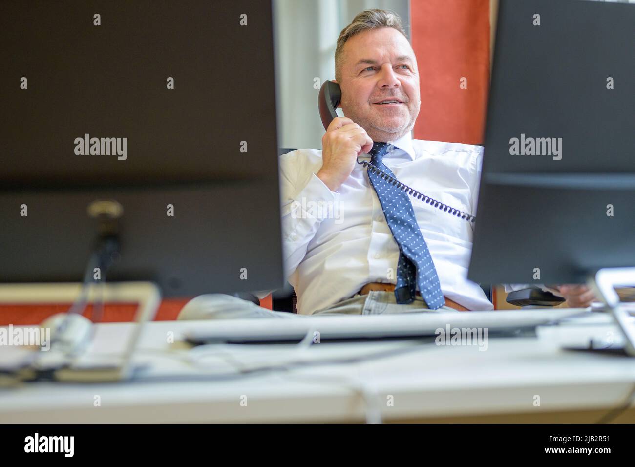 Selbstbewusster Geschäftsmann, der in einem Büro am Telefon spricht, während er sich entspannt, zurückgeschlupft auf seinem Stuhl sitzend Stockfoto