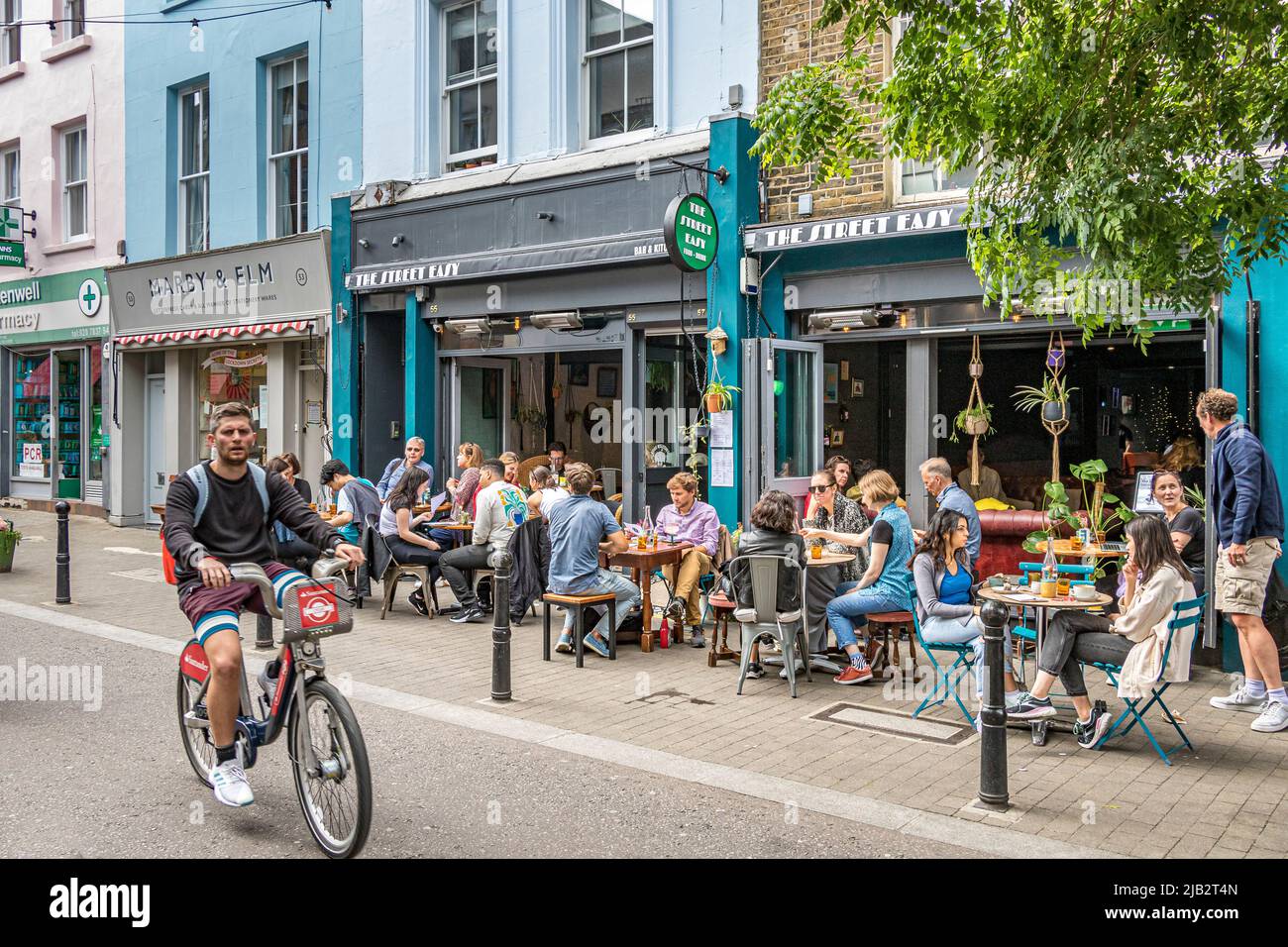 Ein Mann auf dem Fahrrad fährt auf dem Exmouth Market, Clerkenwell, London EC1, an Leuten vorbei, die draußen sitzen und essen und trinken Stockfoto