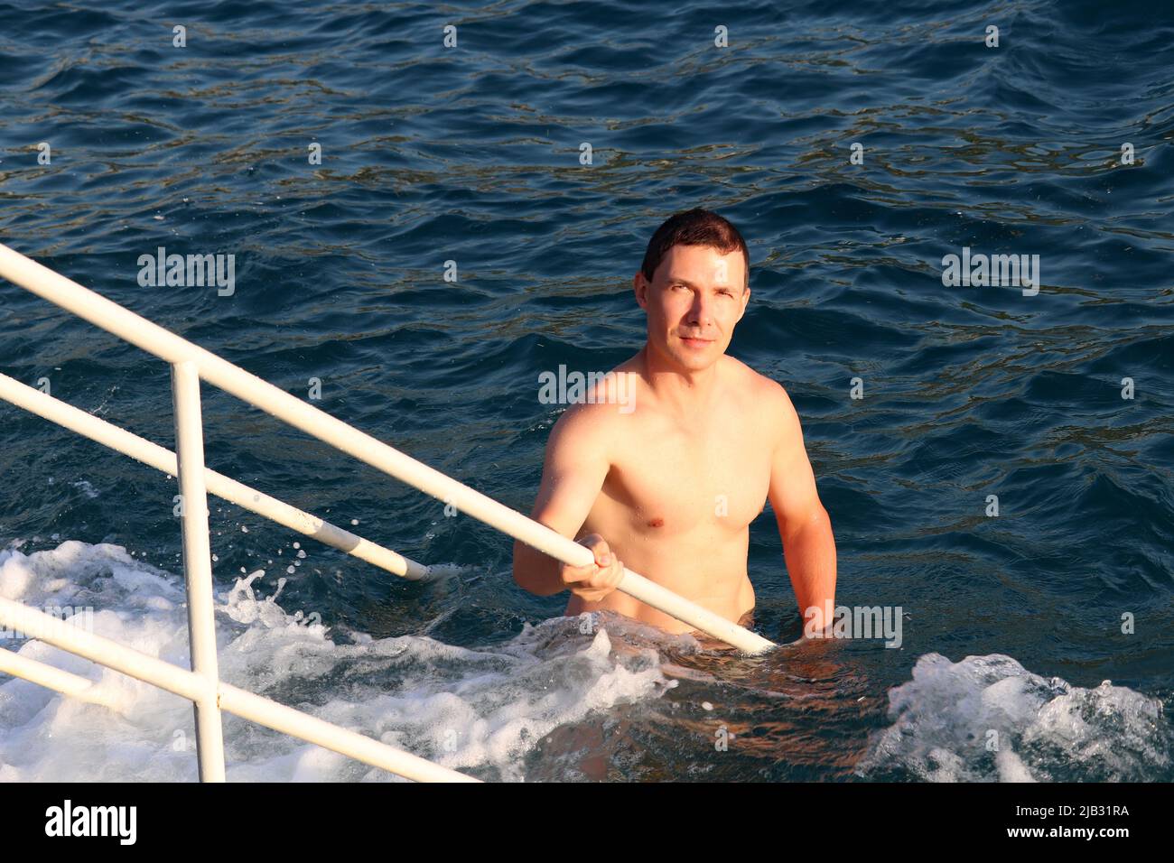 Strandurlaub, Schwimmen im Meer. Porträt eines schönen, muskulösen Mannes, der auf einer Leiter aus blauen Wellen aufsteigt Stockfoto
