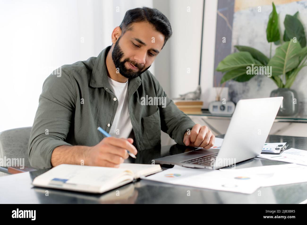 Smart Busy fokussierter Mann arabischer oder indischer Nationalität, erfolgreicher CEO des Unternehmens, Finanzmanager, sitzt an seinem Schreibtisch im modernen Büro, arbeitet mit Dokumenten und Laptop, analysiert, macht Notizen Stockfoto