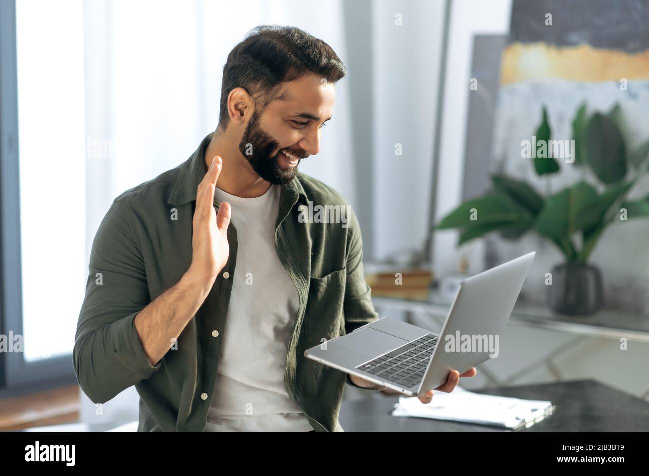 Erfolgreicher arabischer oder indischer Mann, hält und benutzt Laptop, während er im modernen Büro steht, spricht mit Kollegen oder Kunden per Videokonferenz per Laptop, führt ein virtuelles Meeting durch, lächelnd freundlich Stockfoto