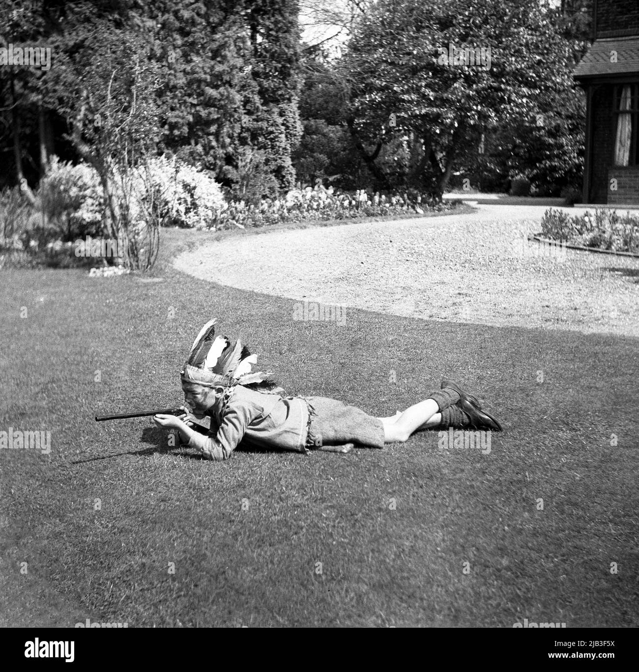 1950s, historisch, draußen im Garten, ein Junge, der spielt, vorgibt, ein gebürtiger oder roter inder zu sein, auf dem Gras liegend, mit seinem Spielzeuggewehr, Federhut und gemaltem Gesicht, England, Großbritannien. In Großbritannien nach dem Krieg war das Spielen von Cowboys und indianern ein beliebtes Spiel für junge Kinder. Stockfoto