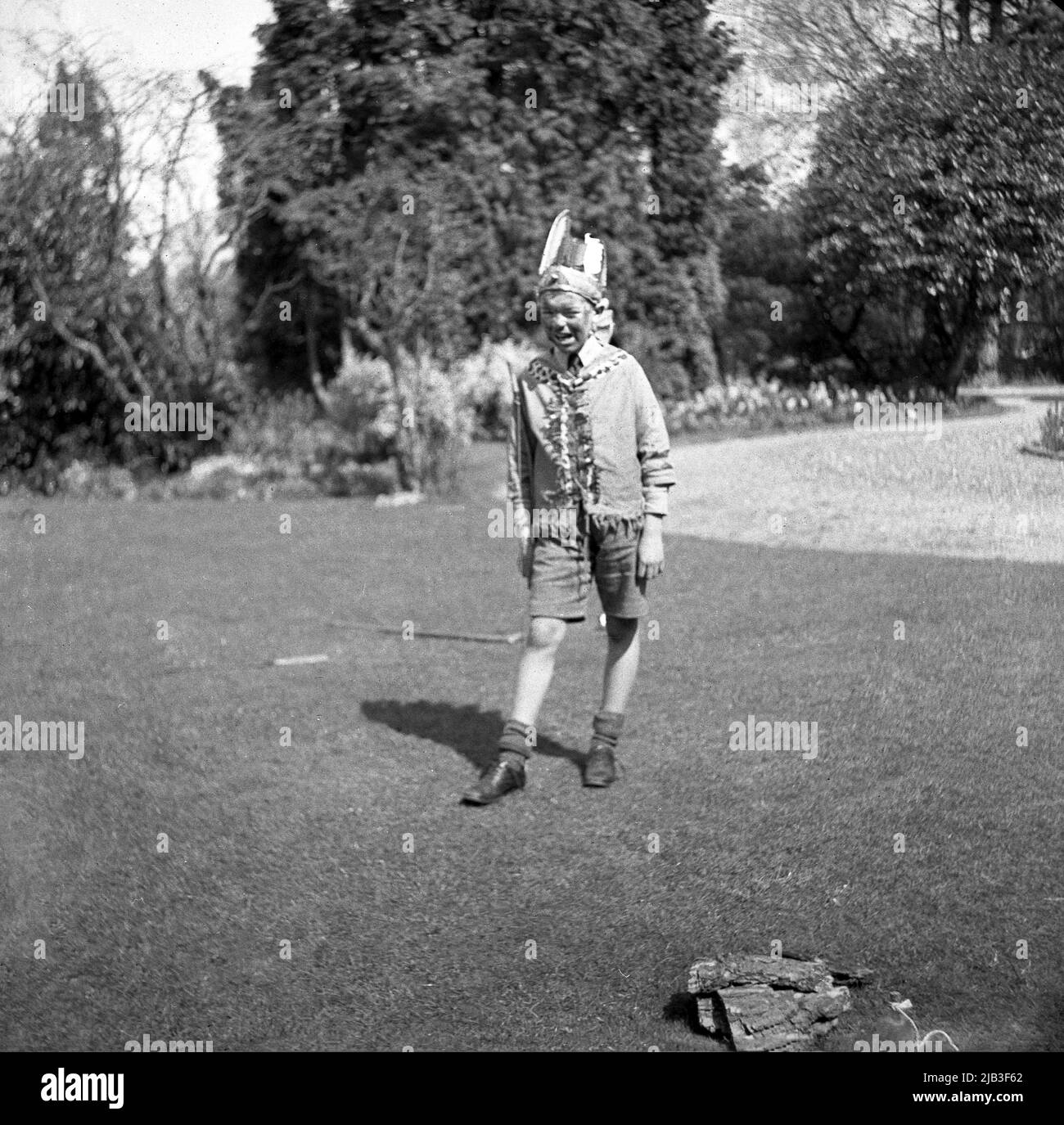 1950s, historisch, draußen im Garten, ein Junge, der auf dem Gras steht, spielt, vorgibt, gebürtig oder ein roter inder zu sein, mit einer Kostümjacke, einer Schleife auf der Schulter, einem Federhut und einem gemalten Gesicht, England, Großbritannien. In Großbritannien nach dem Krieg war das Spielen von Cowboys und indianern ein beliebtes Spiel für junge Kinder. Stockfoto