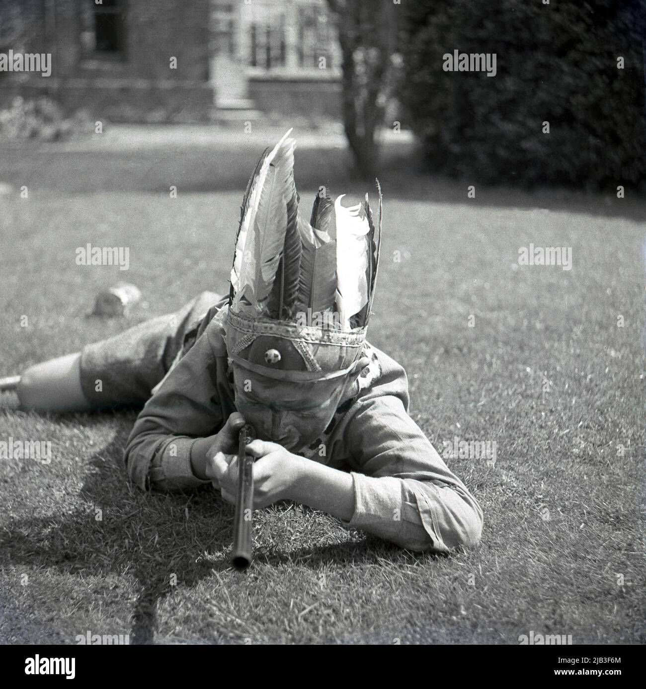 1950s, historisch, draußen im Garten, ein Junge, der spielt, vorgibt, ein gebürtiger oder roter inder zu sein, auf dem Gras liegend, mit seinem Spielzeuggewehr, Federhut und gemaltem Gesicht, England, Großbritannien. In Großbritannien nach dem Krieg war das Spielen von Cowboys und indianern ein beliebtes Spiel für junge Kinder. Stockfoto