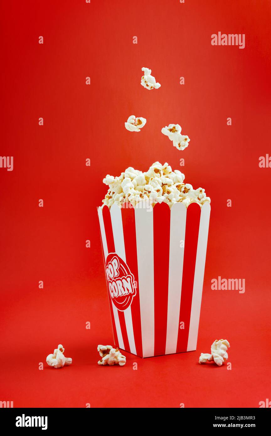 Fallendes Popcorn in einer gestreiften Kartonschachtel auf rotem Hintergrund, vertikale Ansicht. Popcorn Levitation. Kinozeit. Stockfoto