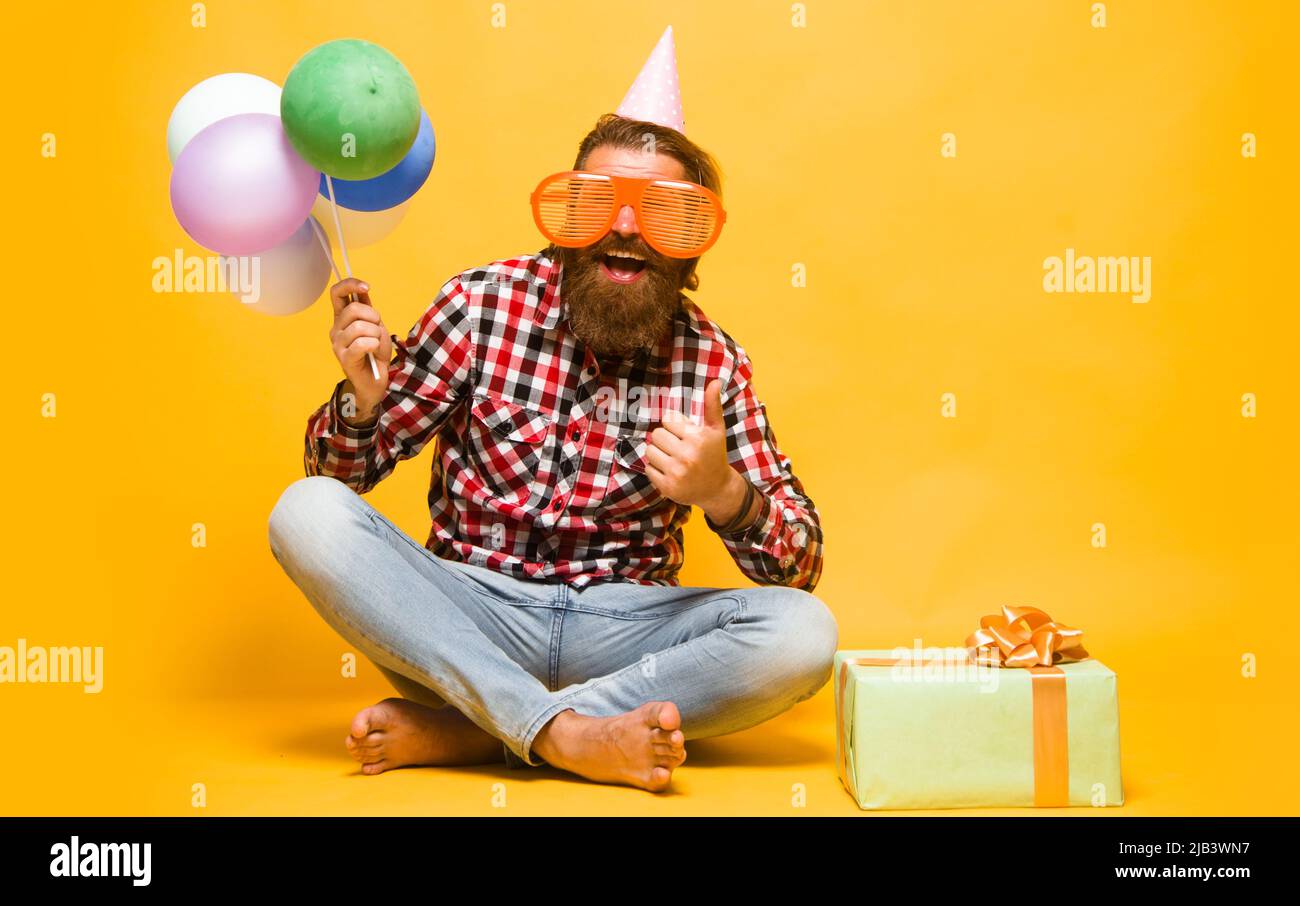 Glücklicher bärtiger Mann im Geburtstagshut und lustige Partybrille mit Ballons und Geschenk zeigt Daumen nach oben. Stockfoto