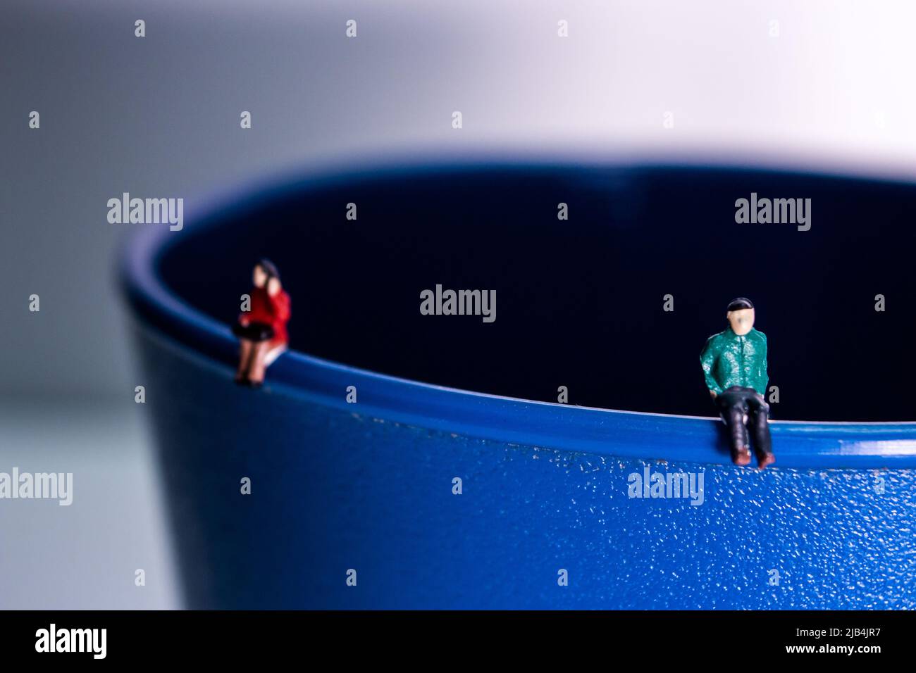 Ein Miniatur-Mann in grünem Outfit sitzt auf dem Rand des blauen Plastikbechers. Der Mann hält Abstand von der Frau. Stockfoto
