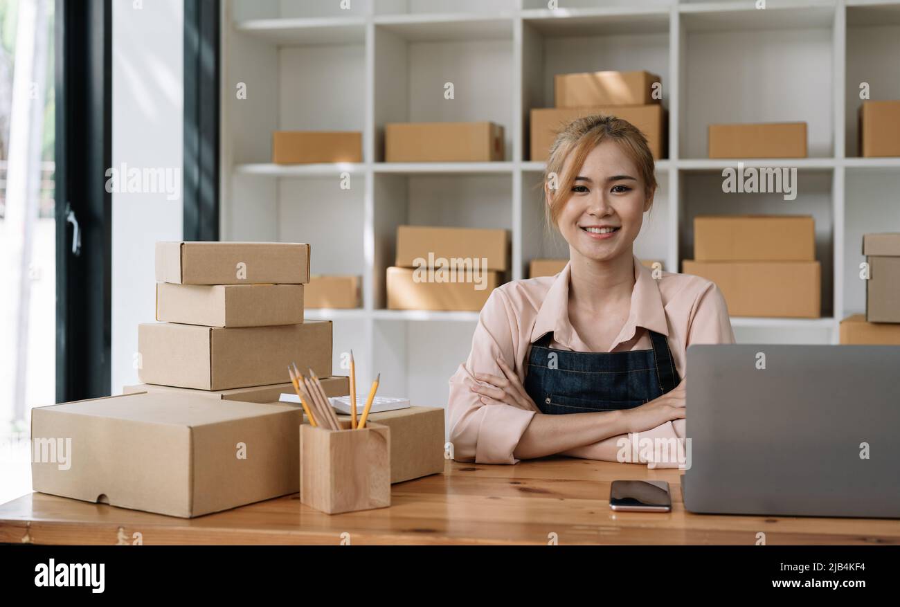 Porträt einer vertraulichen asiatischen Frau Kellnerin Schürze sitzt bei ihr zu Hause mit Pappe für den Versand Online-Shopping Kleinunternehmen Inhaber Konzept. Stockfoto