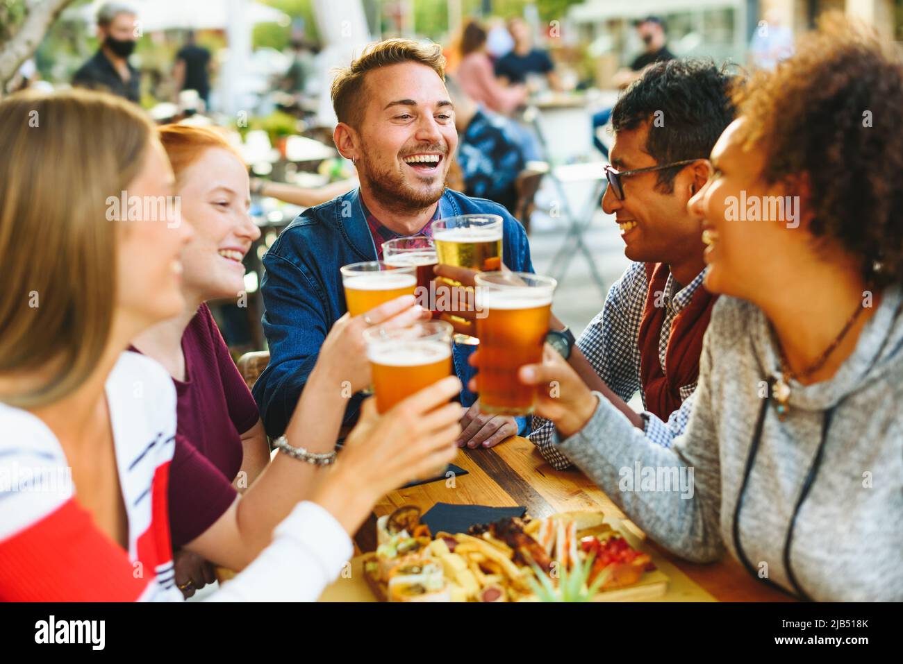 Eine Gruppe von begeisterten multirassischen Freunden, die sich gegenseitig angucken, während sie mit Biergläsern am Tisch mit Essen auf der Terrasse des Pubs jubeln Stockfoto