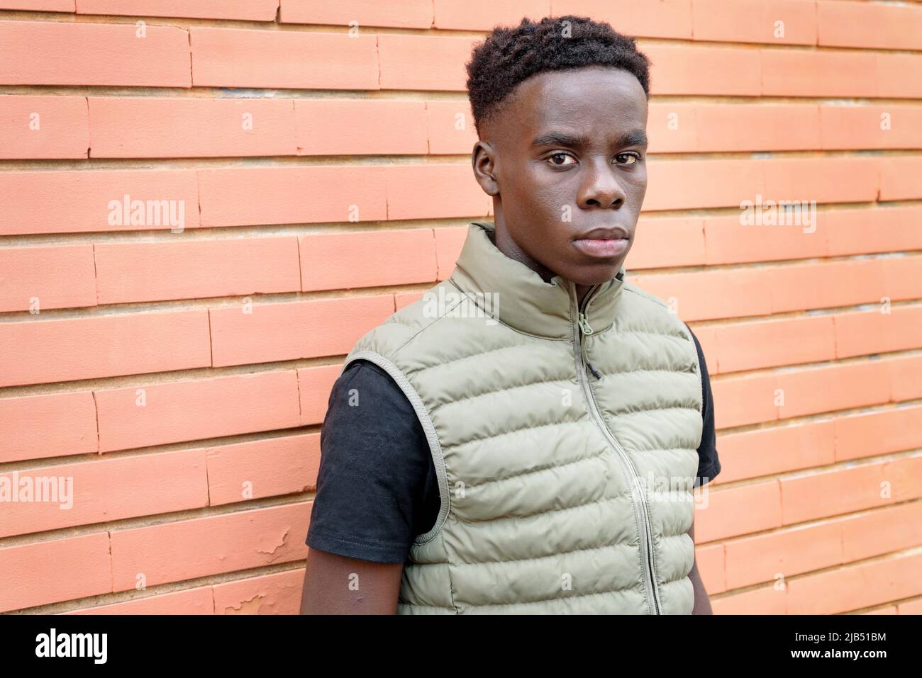Ernsthafter afrikanischer Mann in Weste, der in der Nähe einer orangefarbenen Ziegelsteinmauer auf der Stadtstraße die Kamera ansah Stockfoto