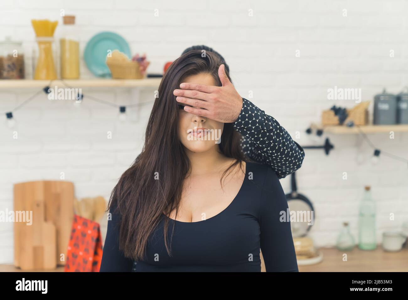 Nicht erkennbarer Mann, der seine kaukasische langhaarige Freundin mit einem Geschenk überraschte, indem er ihre Augen mit seiner Hand bedeckte. Küchenausstattung. Hochwertige Fotos Stockfoto