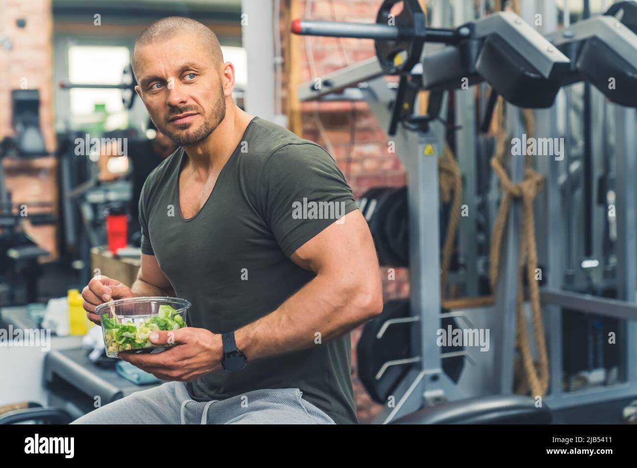 Die Bedeutung der Ernährung in den Fettabbau. Muskulöser kahlköpfiger bärtiger Mann, der einen gesunden Salat isst, während er auf einer Fitnessbank sitzt. Hochwertige Fotos Stockfoto