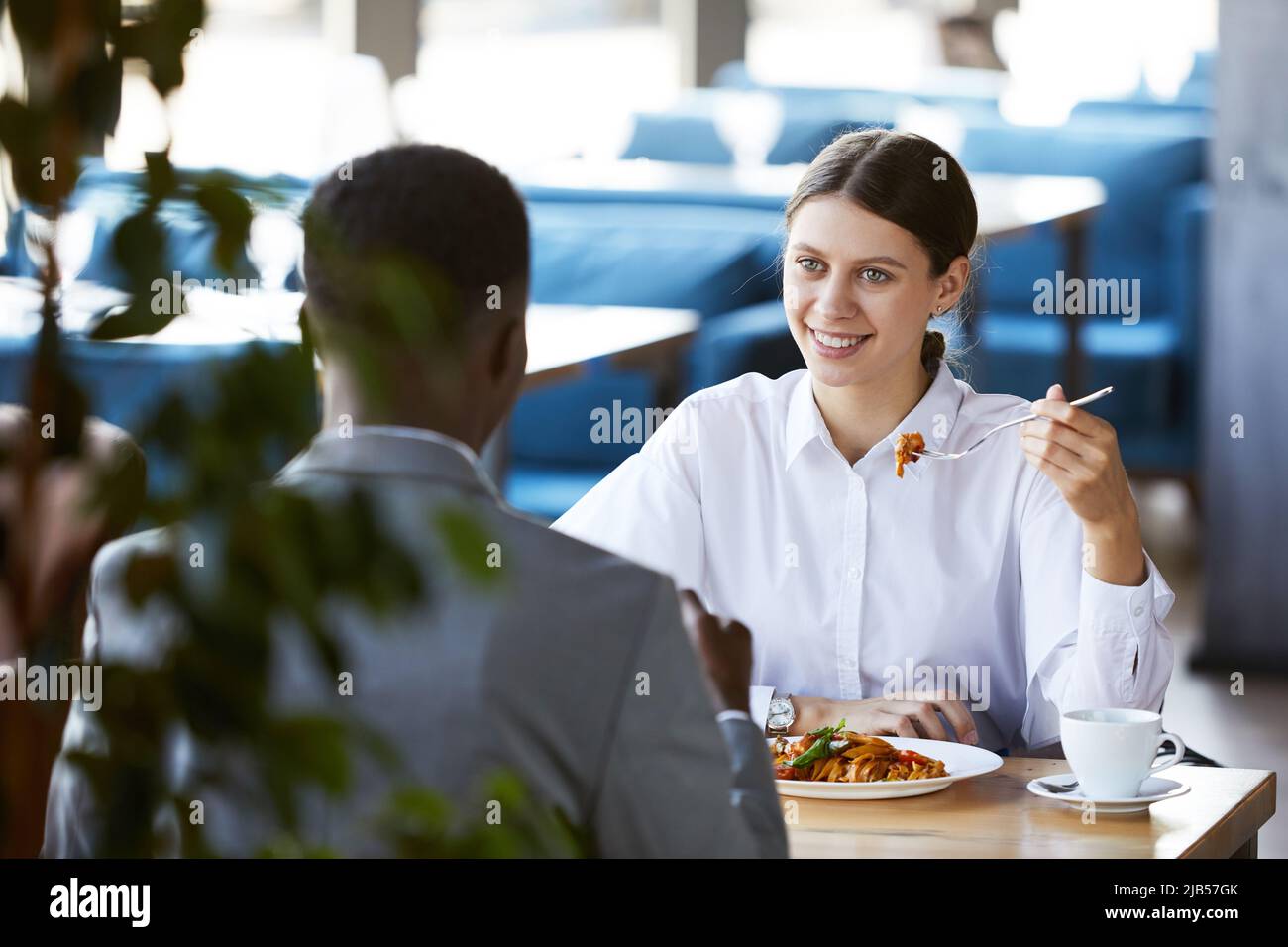 Positive aufgeregte junge Geschäftsfrau im weißen Hemd, die am Tisch sitzt und Pasta isst, während sie sich mit der schwarzen Führungskraft im Café trifft Stockfoto