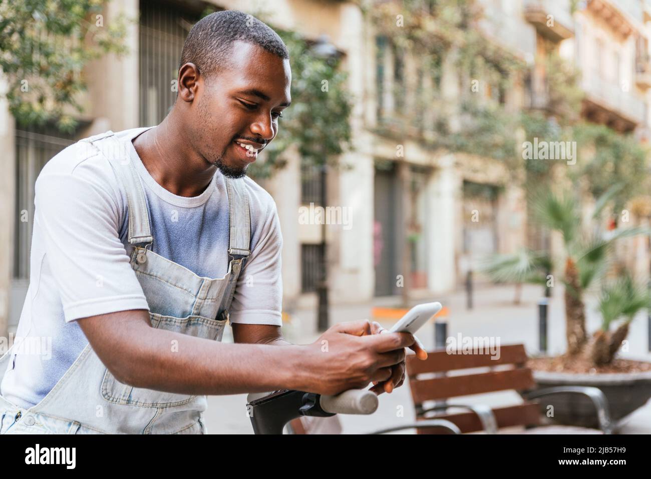Positive junge schwarze Männer Millennial mit dunklen Haaren und Bart lächeln und Messaging auf Handy, während sich auf Kick Scooter auf der Straße der Stadt Stockfoto